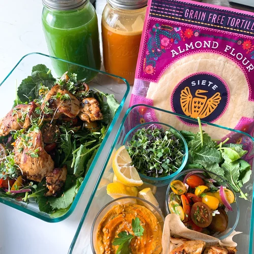 Salad with greens, cherry tomatoes, lemon slices, and a bowl of orange-colored dip; jars of green and orange drinks, and almond flour package in background.