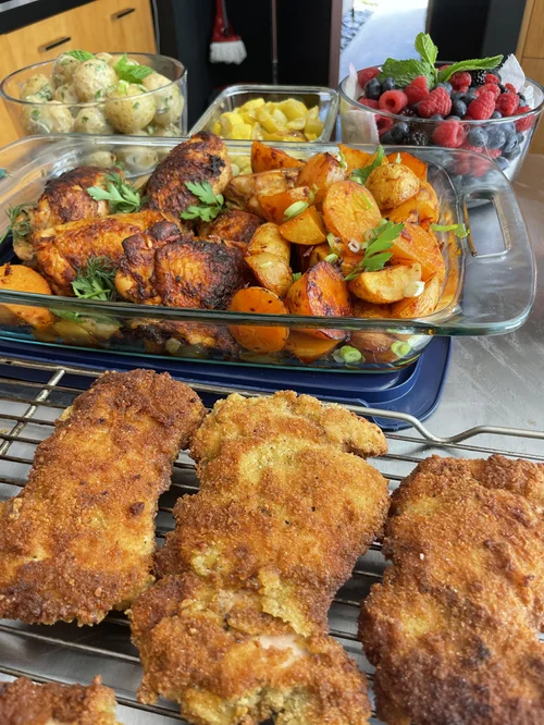 Fried chicken pieces on a cooling rack, with a glass dish of roasted vegetables and bowls of fruit in the background.