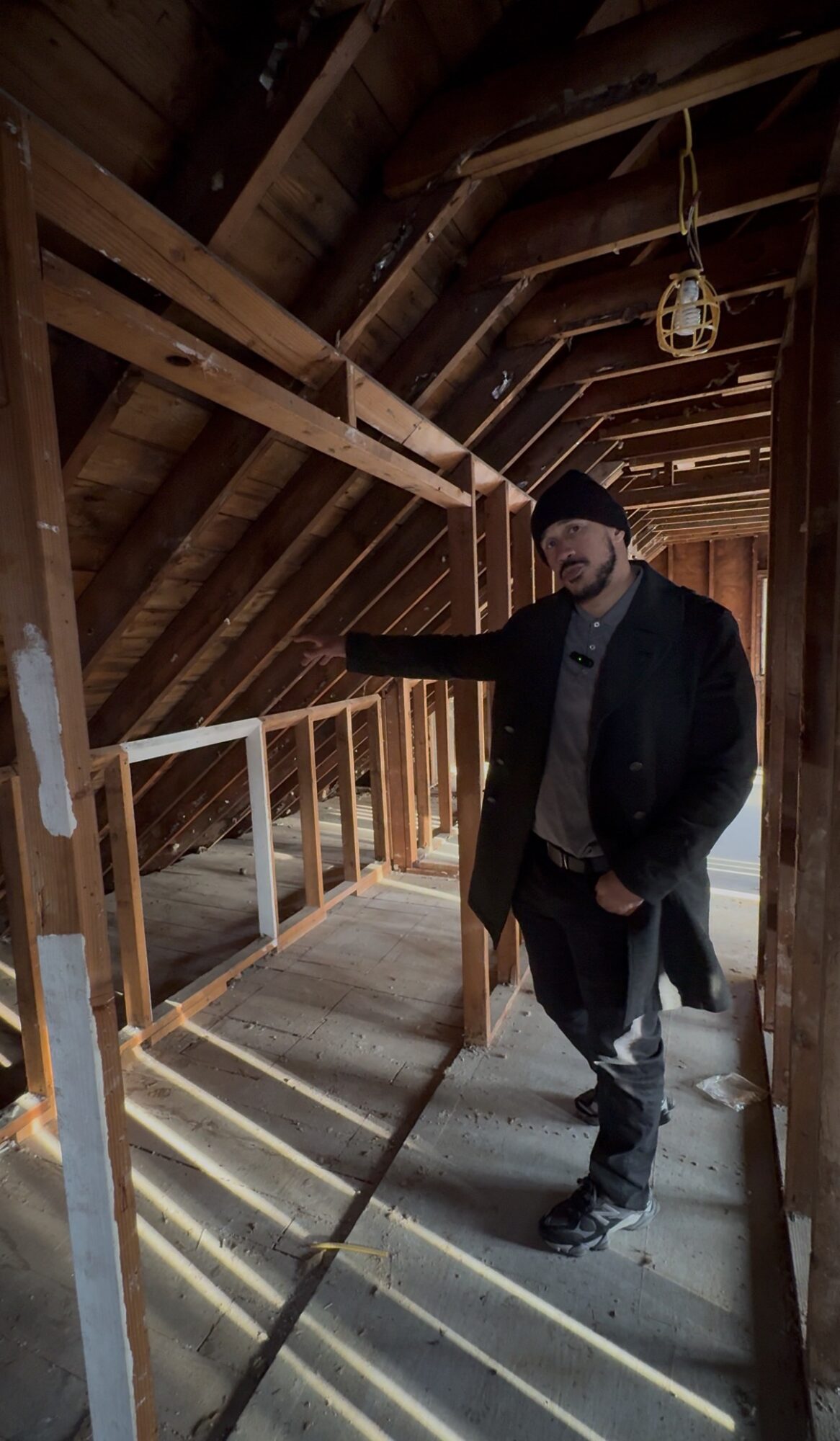Man in dark clothing and beanie standing inside an unfinished wooden attic, pointing at the wall with sunlight streaming through gaps.
