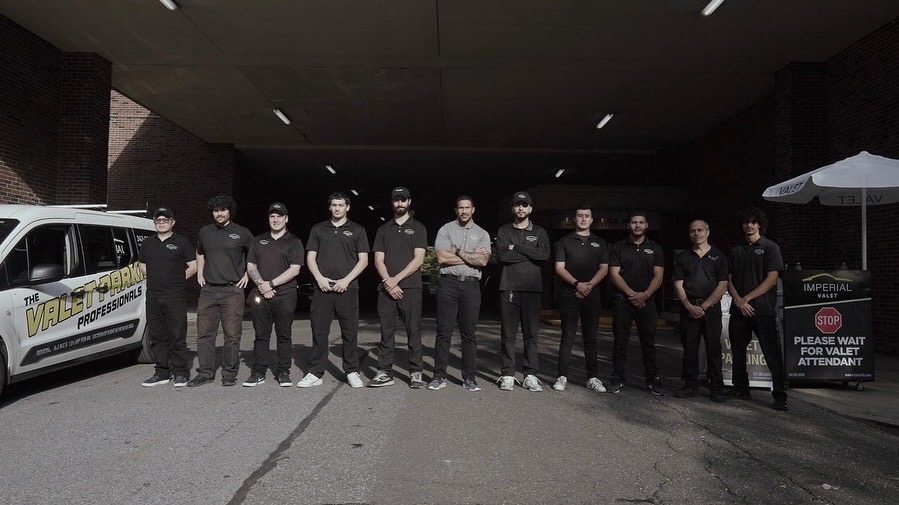 Group of ten people standing outdoors under a dark overpass, wearing black shirts and masks, with a white vehicle nearby.