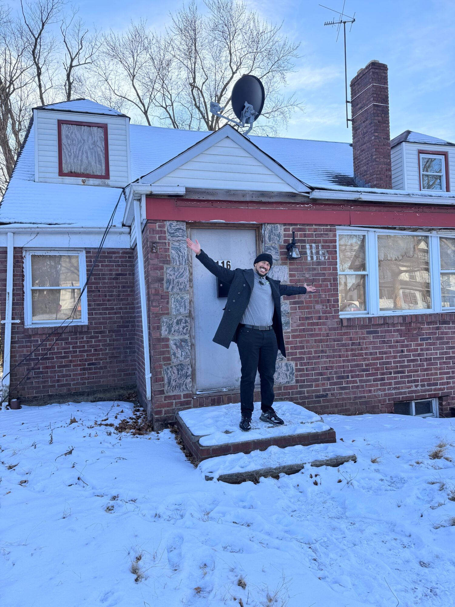 Person standing on snow-covered ground outside brick house, arms outstretched, winter scene, trees in background.