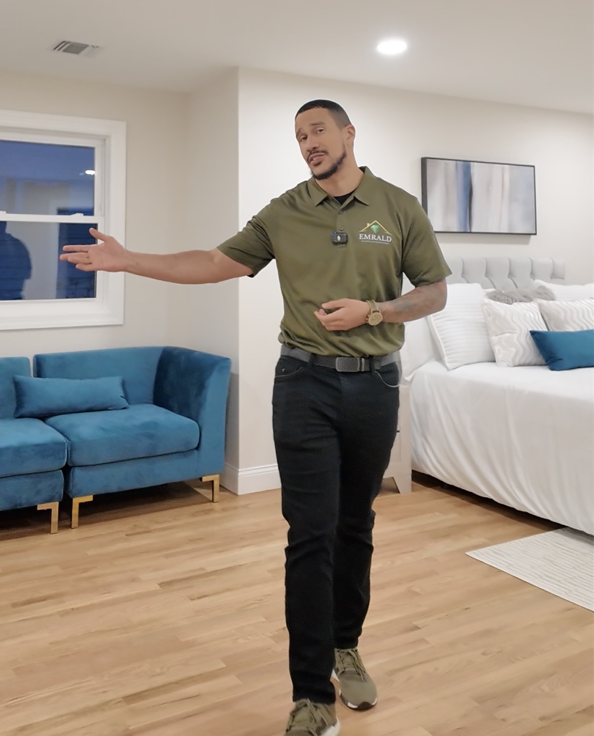 Person in uniform gesturing with hand in a modern bedroom with blue sofa and white bed, hardwood floor, and window.