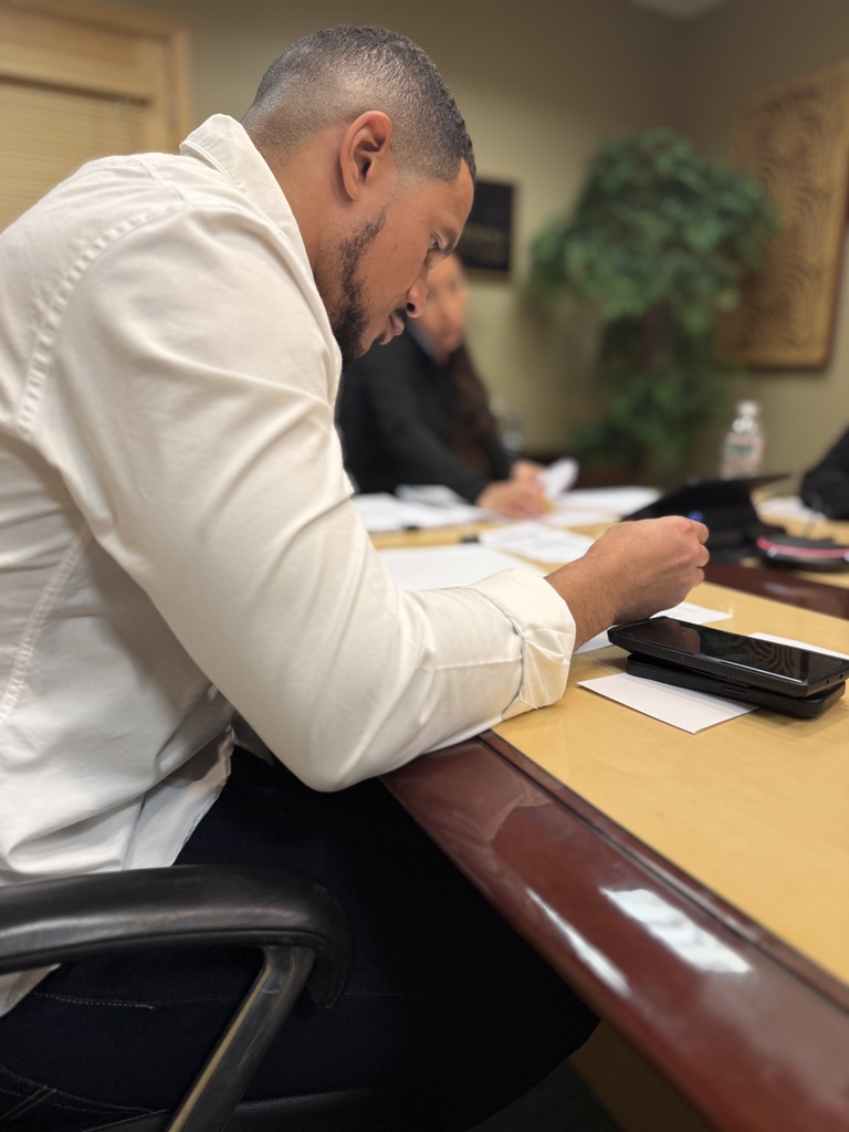Man in white shirt writing at a conference table with papers and a calculator, blurred background with plant and framed picture.