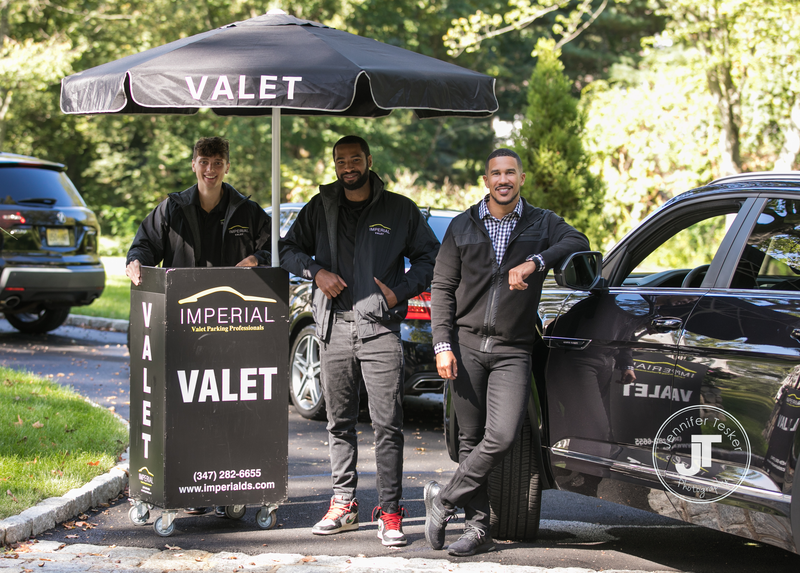 Three men standing near valet stand with umbrella, cars in background, trees, and sunlight, smiling and posing outdoors.
