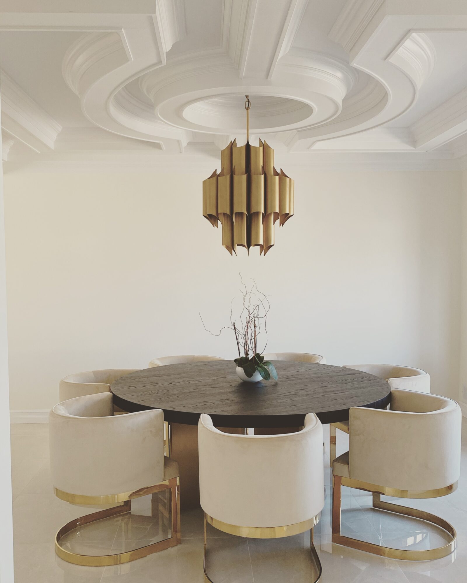 Round dining table with six cream-colored chairs in a room with ornate ceiling molding and a gold chandelier.