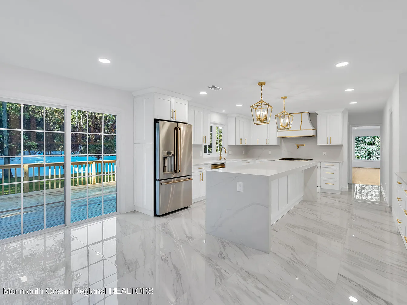 Bright, modern kitchen with white cabinets, marble floors, large island, stainless steel refrigerator, and sliding glass doors to outdoor area.