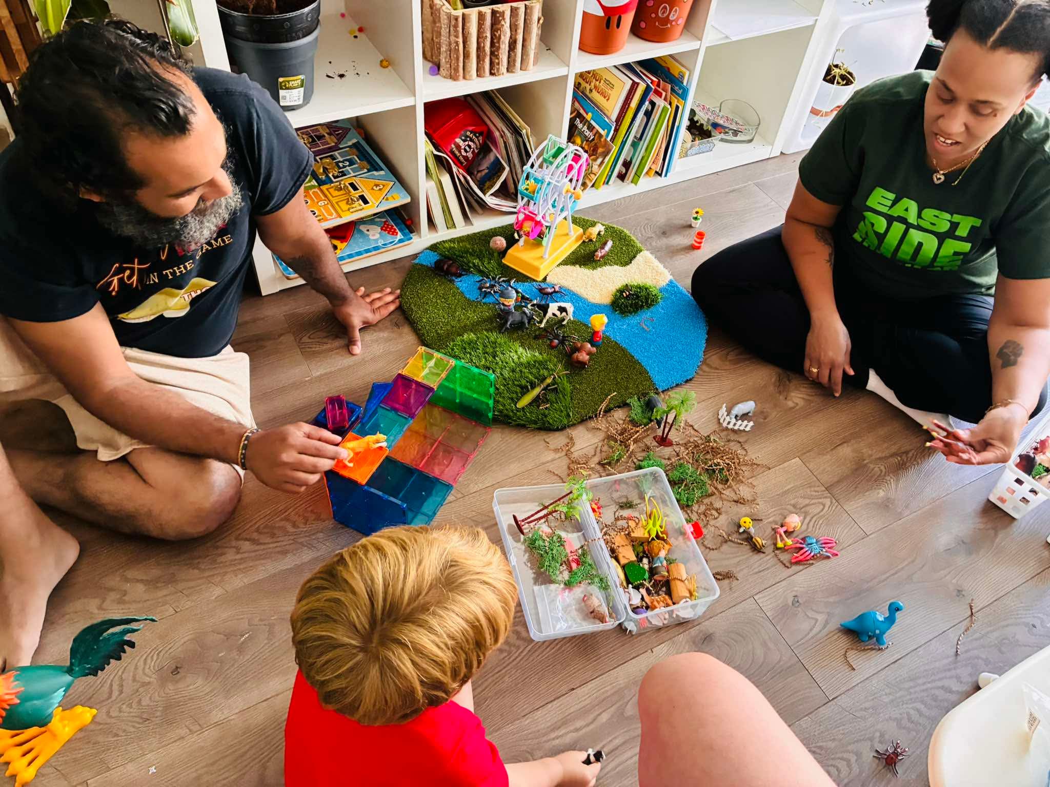 Two women and a child sit on the floor playing with colorful toys and a small model landscape, surrounded by books and toys.