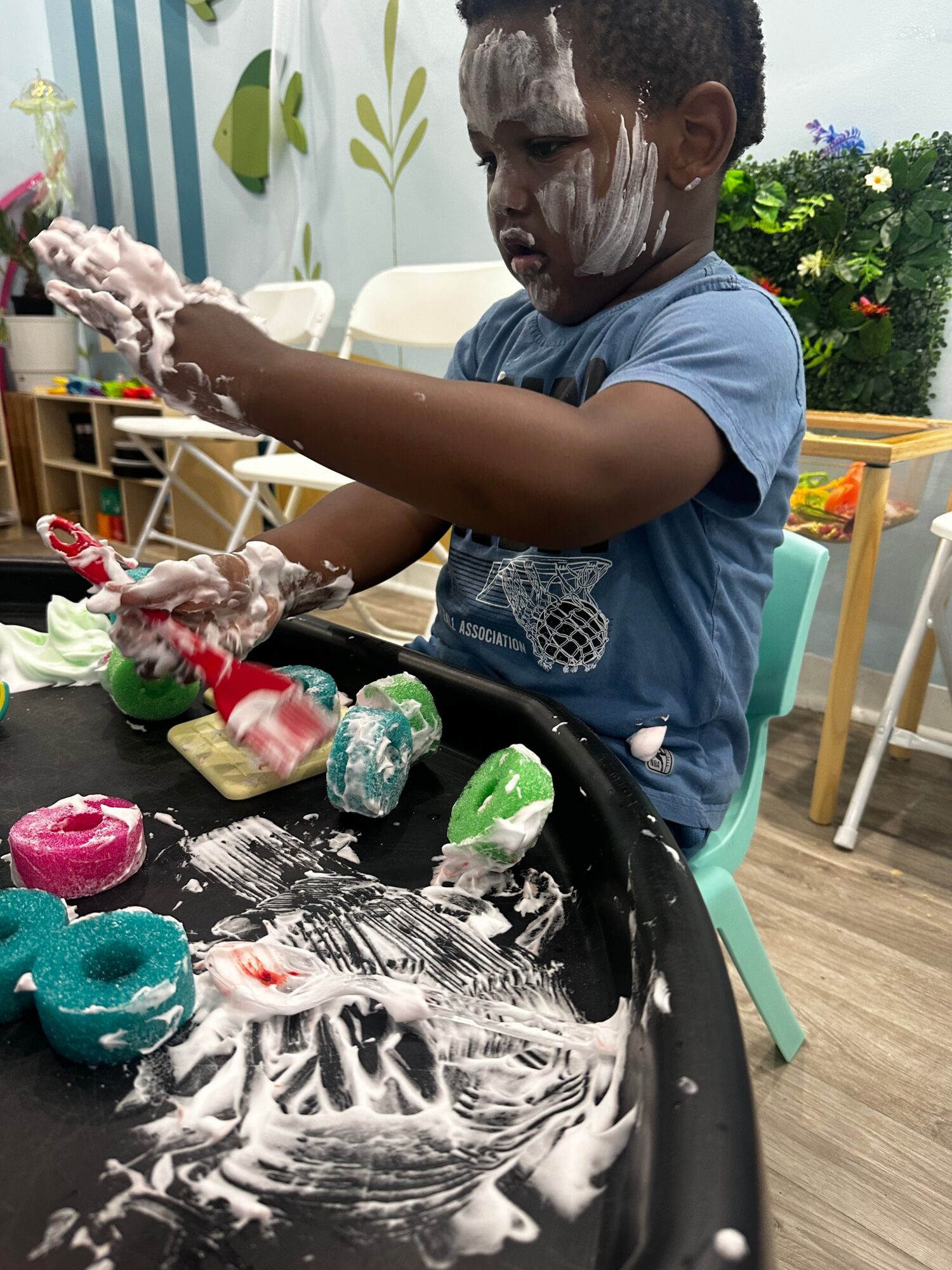 Child playing with soap and colorful toys at a table outdoors, with plants and furniture in background.