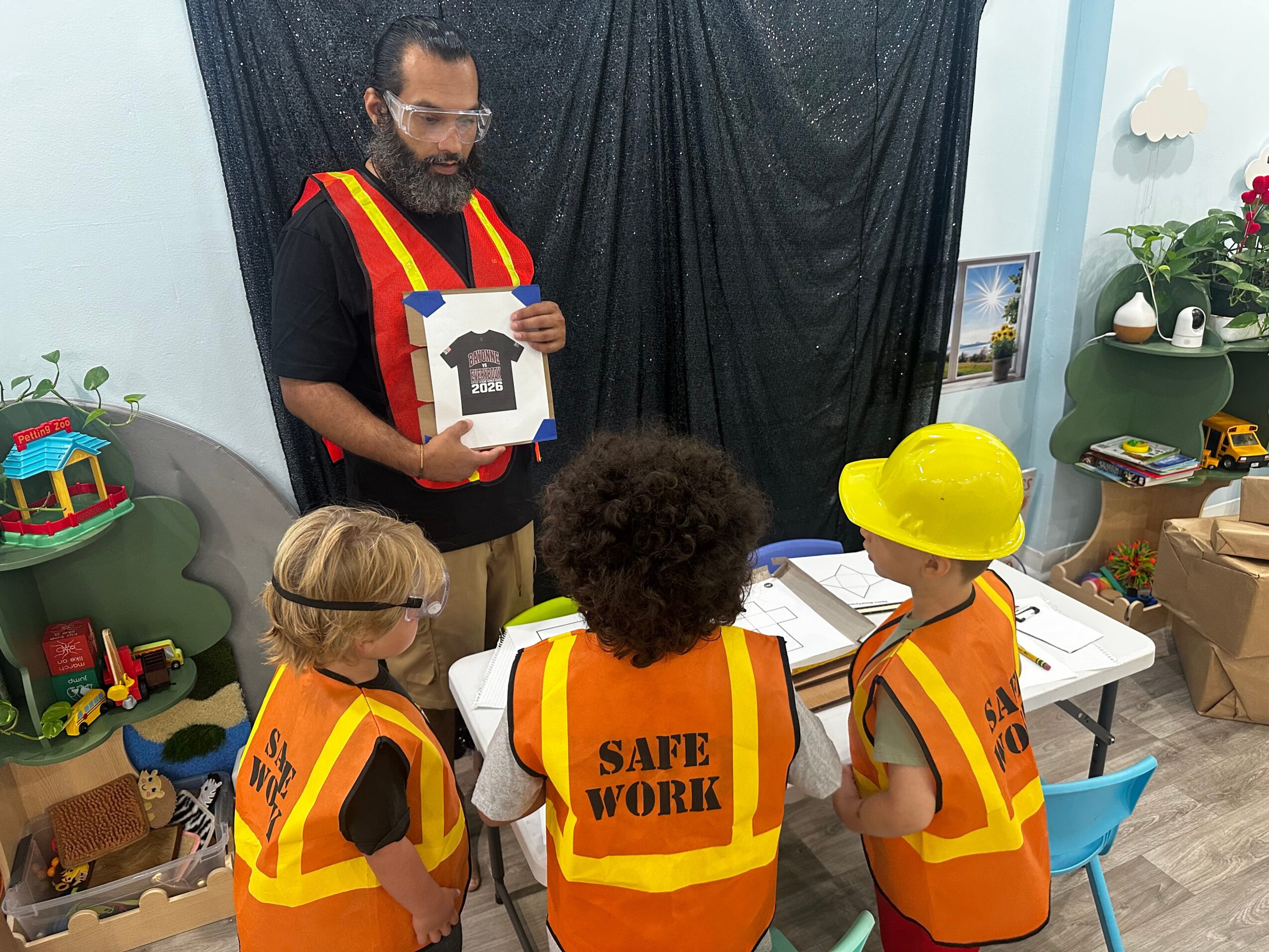 Adult showing children in safety vests a picture of a t-shirt, in a room with toys and plants.
