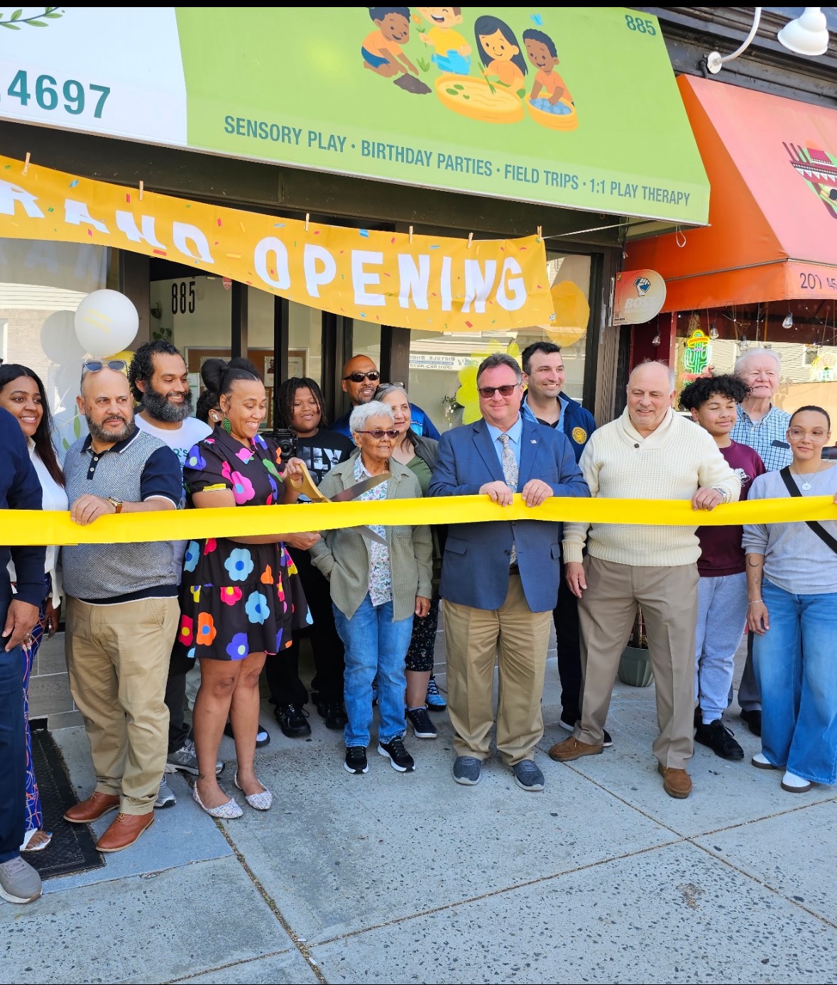 Group of people at a ribbon-cutting event outside a store with a yellow banner and sign, celebrating grand opening.
