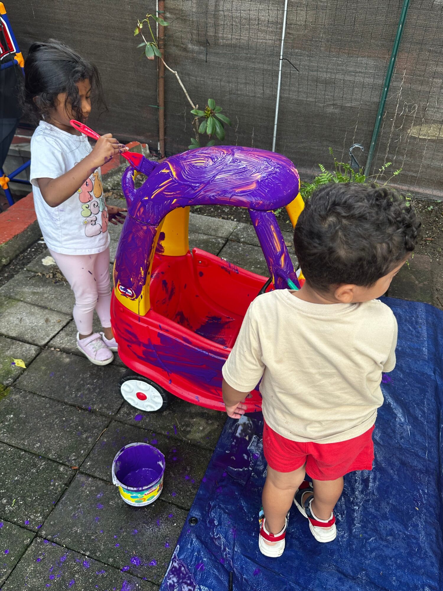 Child and girl playing with a red and purple toy on a wet outdoor surface, with a blue tarp nearby.