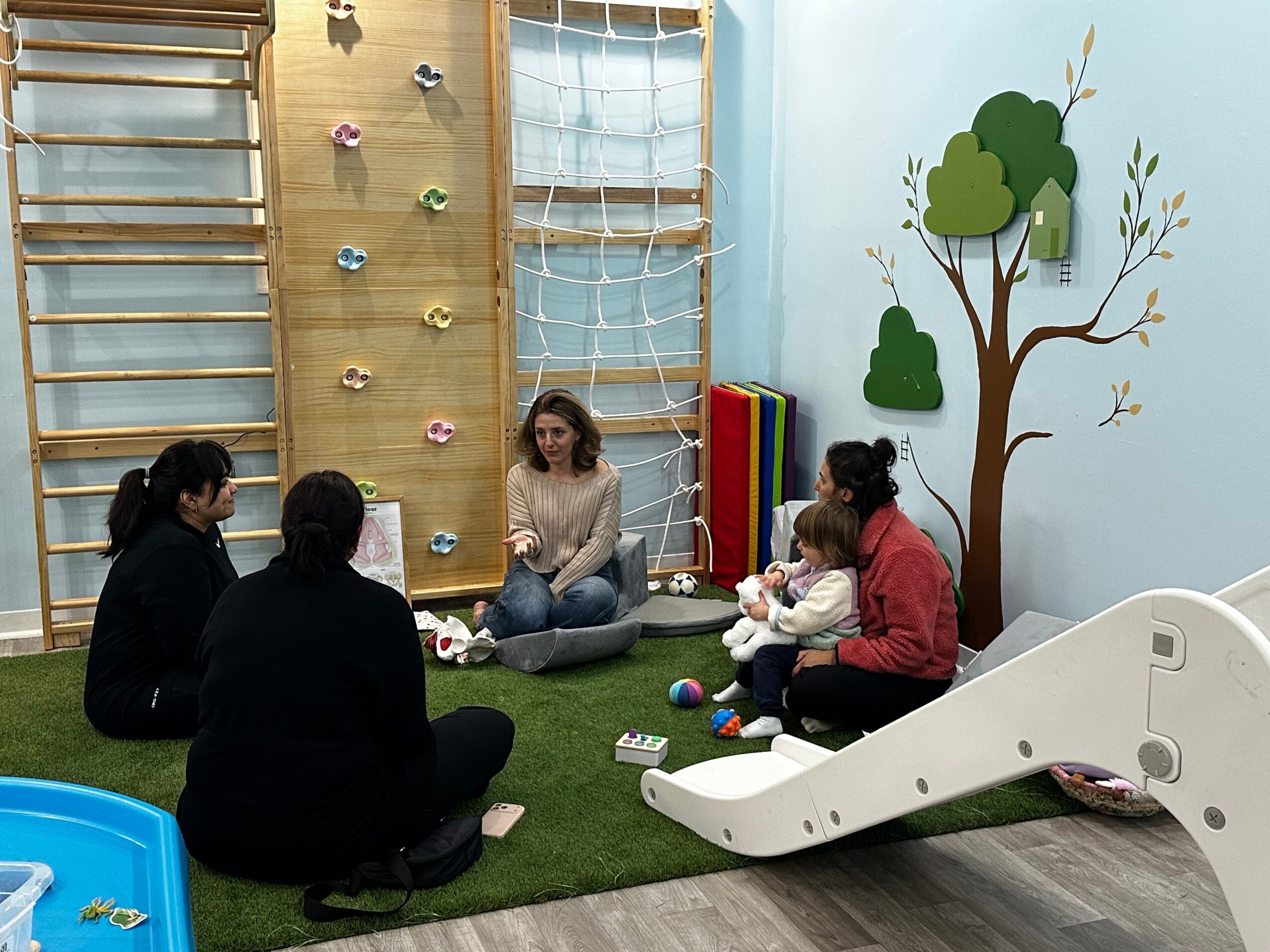 Four women and a child sit on a green carpet in a playroom with climbing walls and a slide, engaging in conversation.