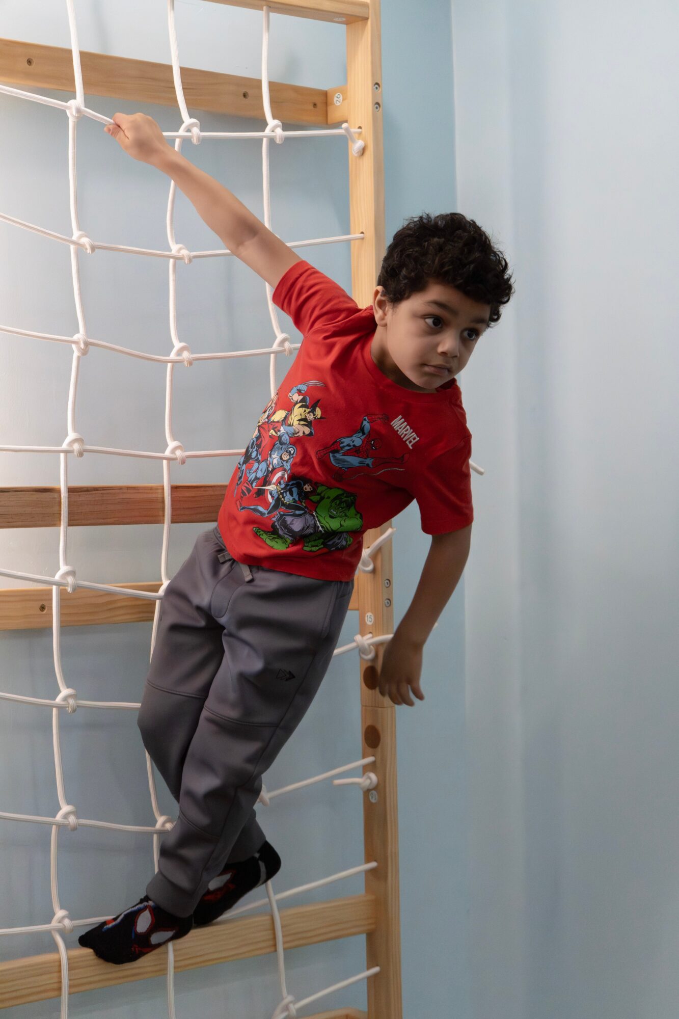 Child climbing on a rope net structure indoors, holding onto the net with one hand, wearing a red shirt and gray pants.