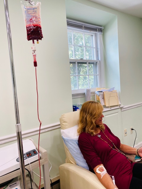 Woman receiving intravenous therapy in a hospital room with a window and medical equipment, sitting in a chair with a bandage on her arm.