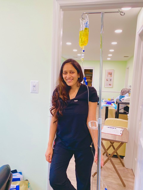 Woman standing in a room with IV pole and medical equipment, smiling, wearing dark scrubs, with a bandage on her arm.