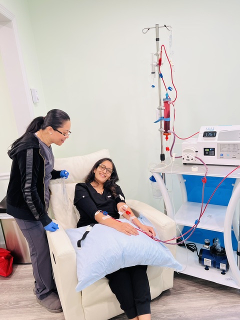 A woman is lying on a hospital bed connected to medical equipment, with another woman standing beside her, both smiling.