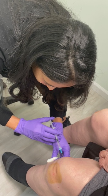 Person with long dark hair wearing purple gloves administering an injection to a person's thigh.