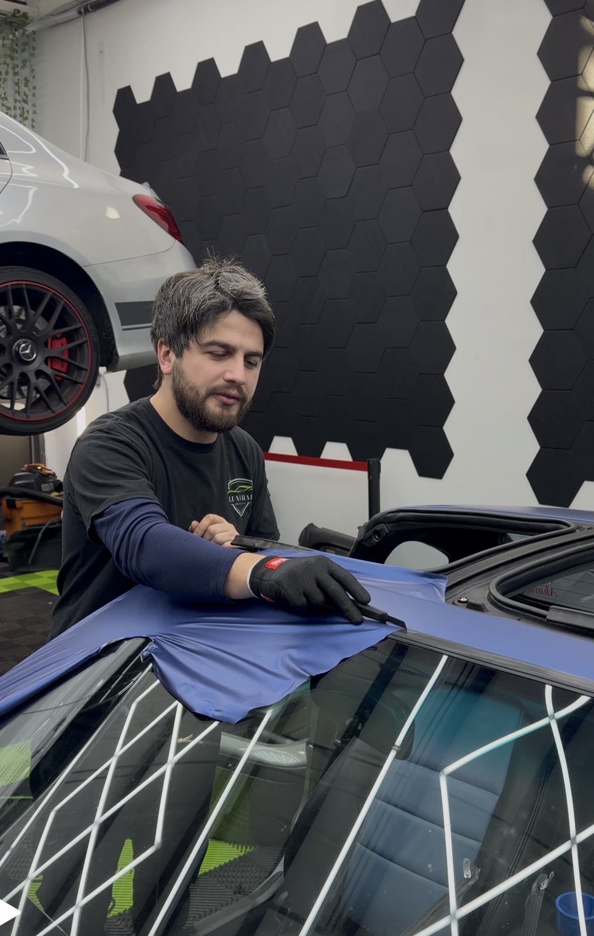 Man working on a car in a garage with black and gray wall panels, a white car, and tools.