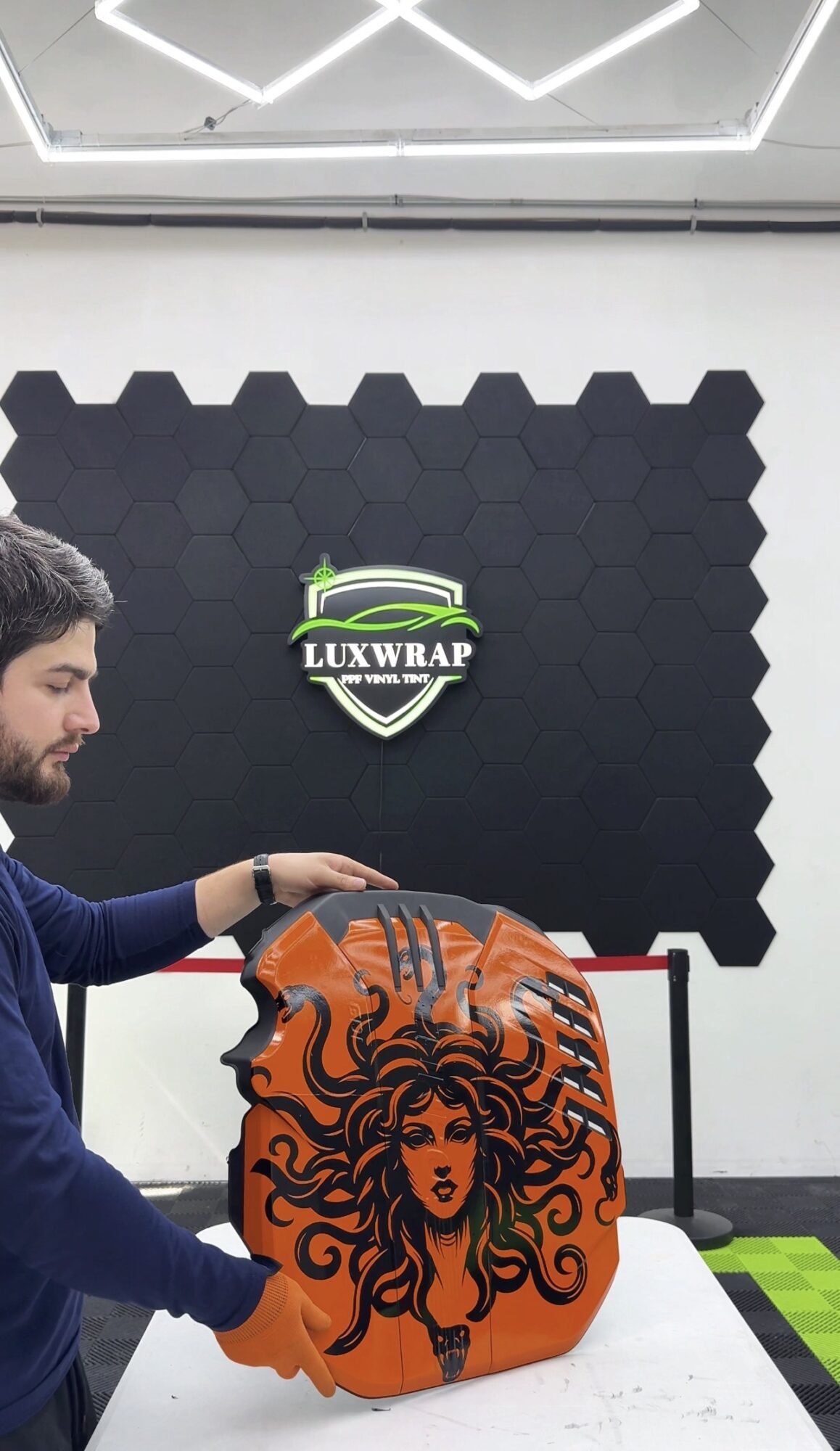 Person with dark hair and beard holding an orange bag with a black artistic face design, in a modern room.