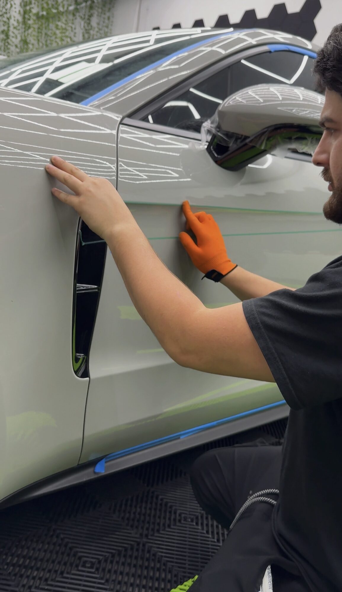 Person in black clothing and orange glove working on a silver car's door, with a side mirror visible.