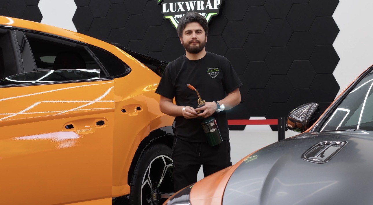 Man holding a bottle standing between two cars, one orange and one gray, in an indoor setting.