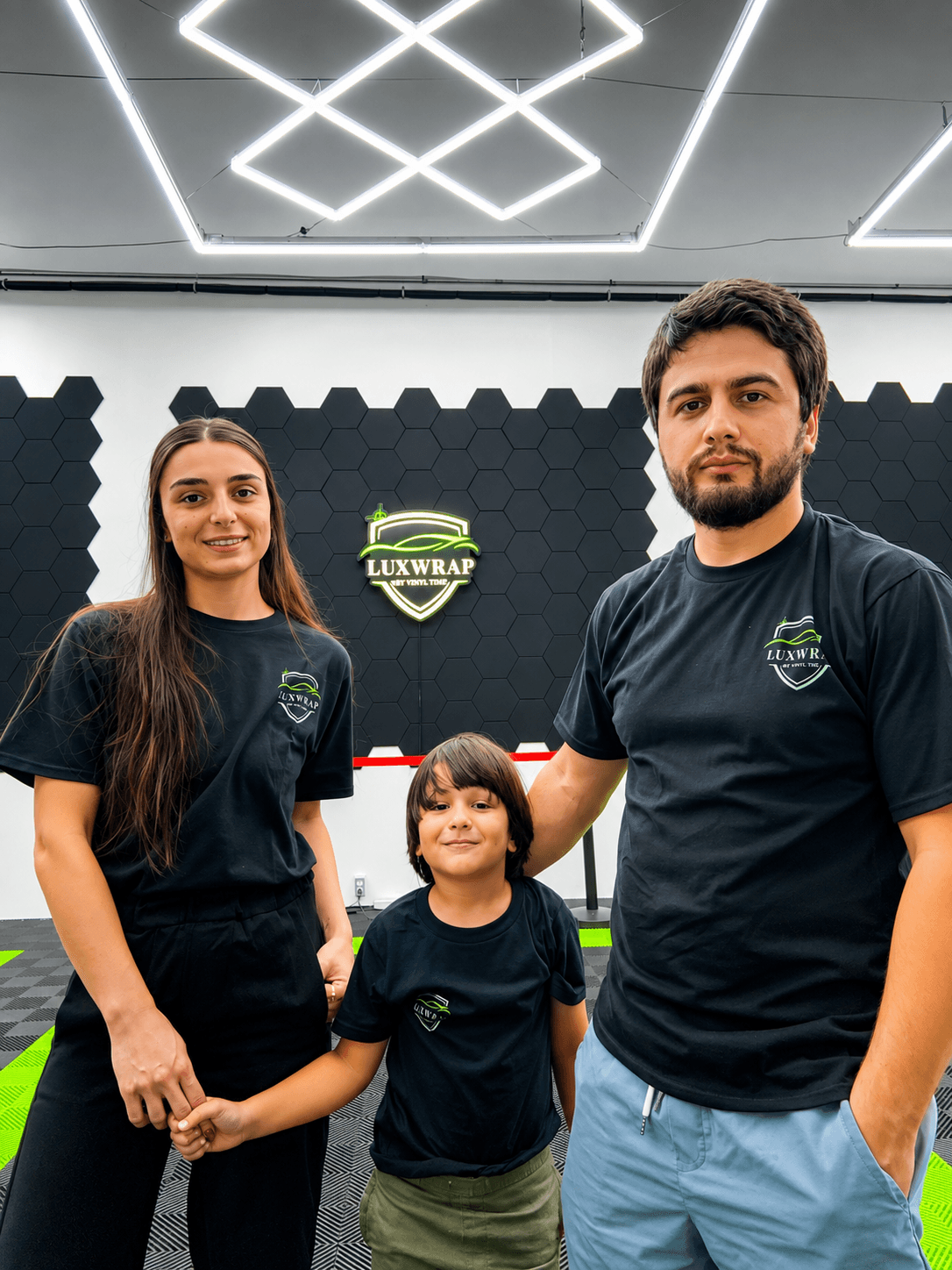 Three people, two adults and one child, stand indoors in front of a black wall with a logo, smiling.