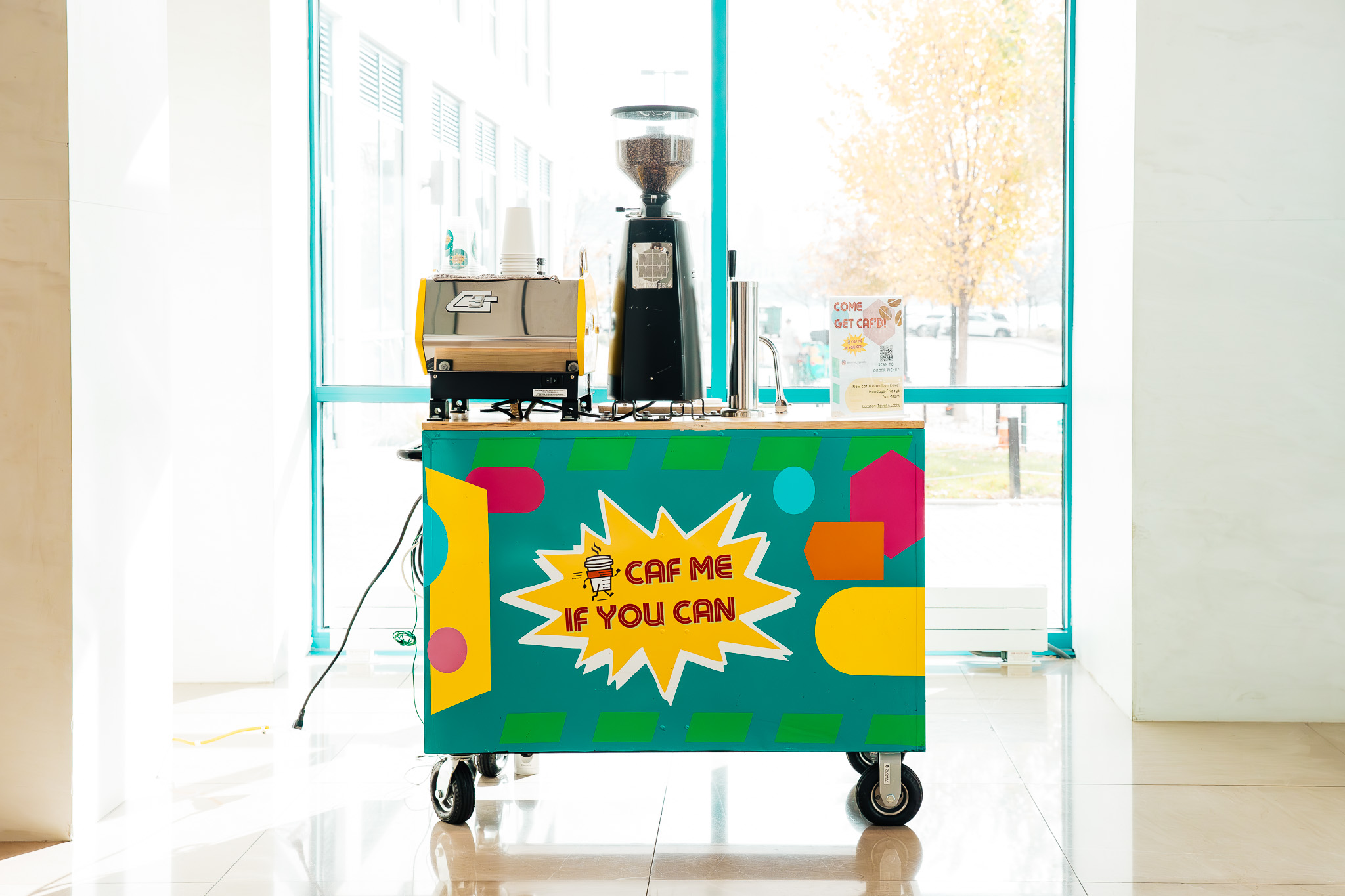 Coffee cart with colorful design and sign, coffee grinder, and equipment inside a bright room with large windows.