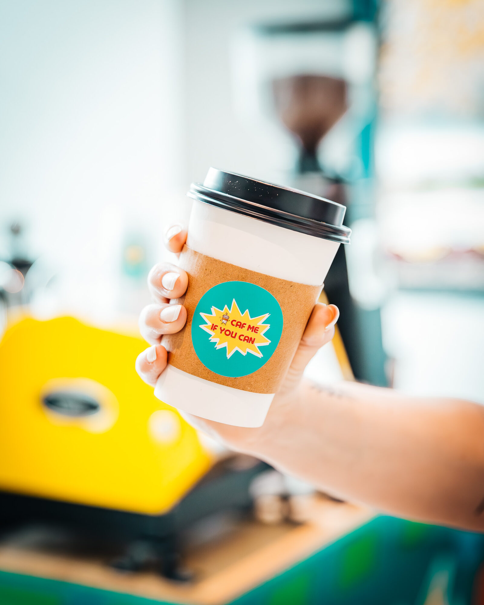 Person holding a disposable coffee cup with a black lid, in a bright indoor setting.