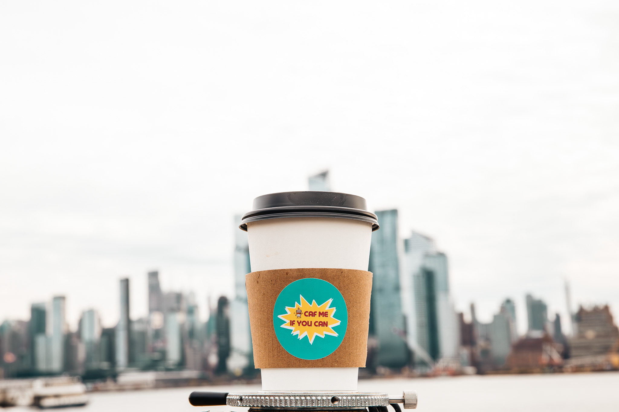 Takeaway coffee cup with city skyline in background, cloudy sky, and a colorful sticker on the cup.