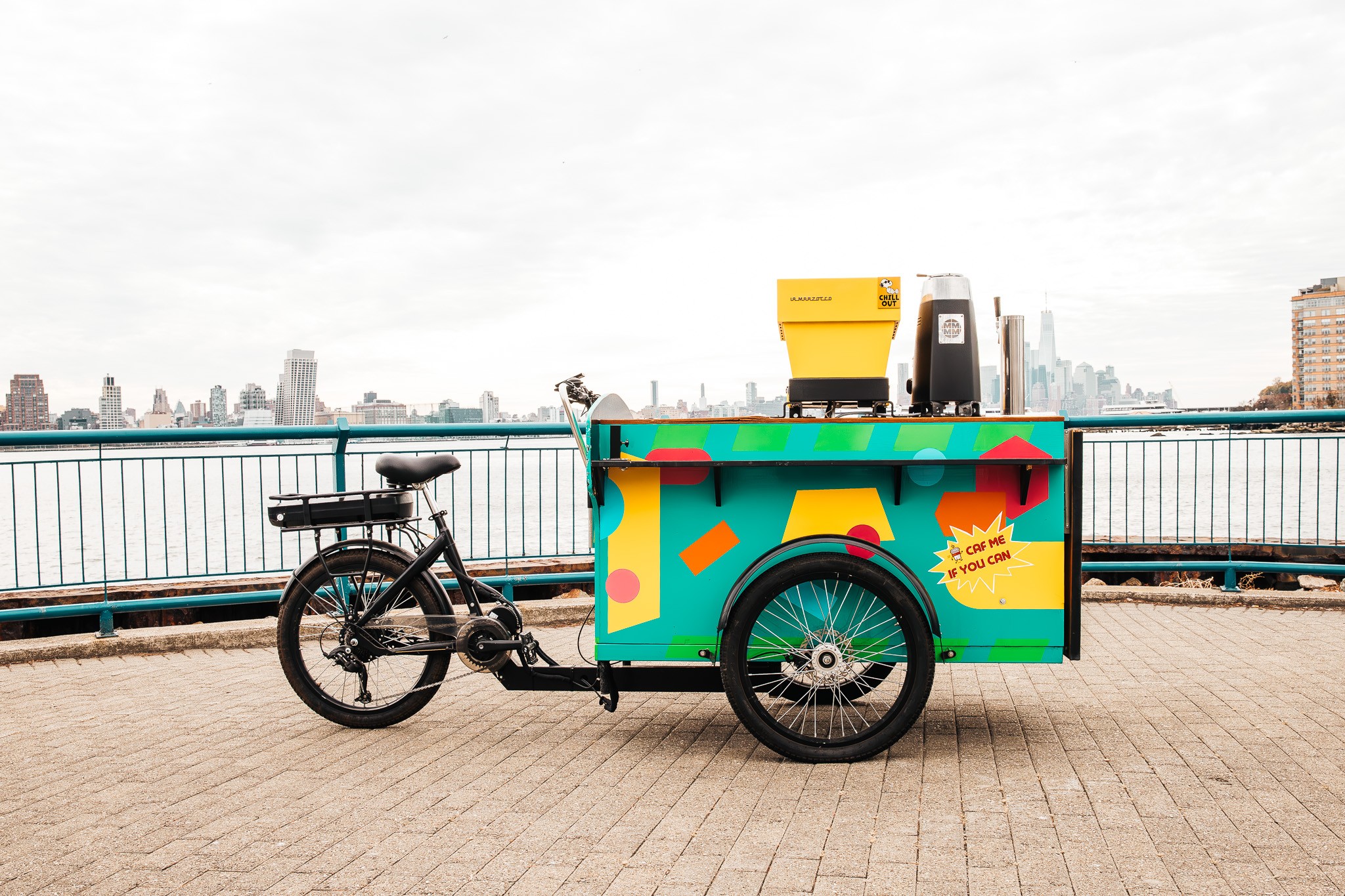 Bike attached to a colorful cart with a yellow box on top, on a waterfront promenade with city skyline in background.