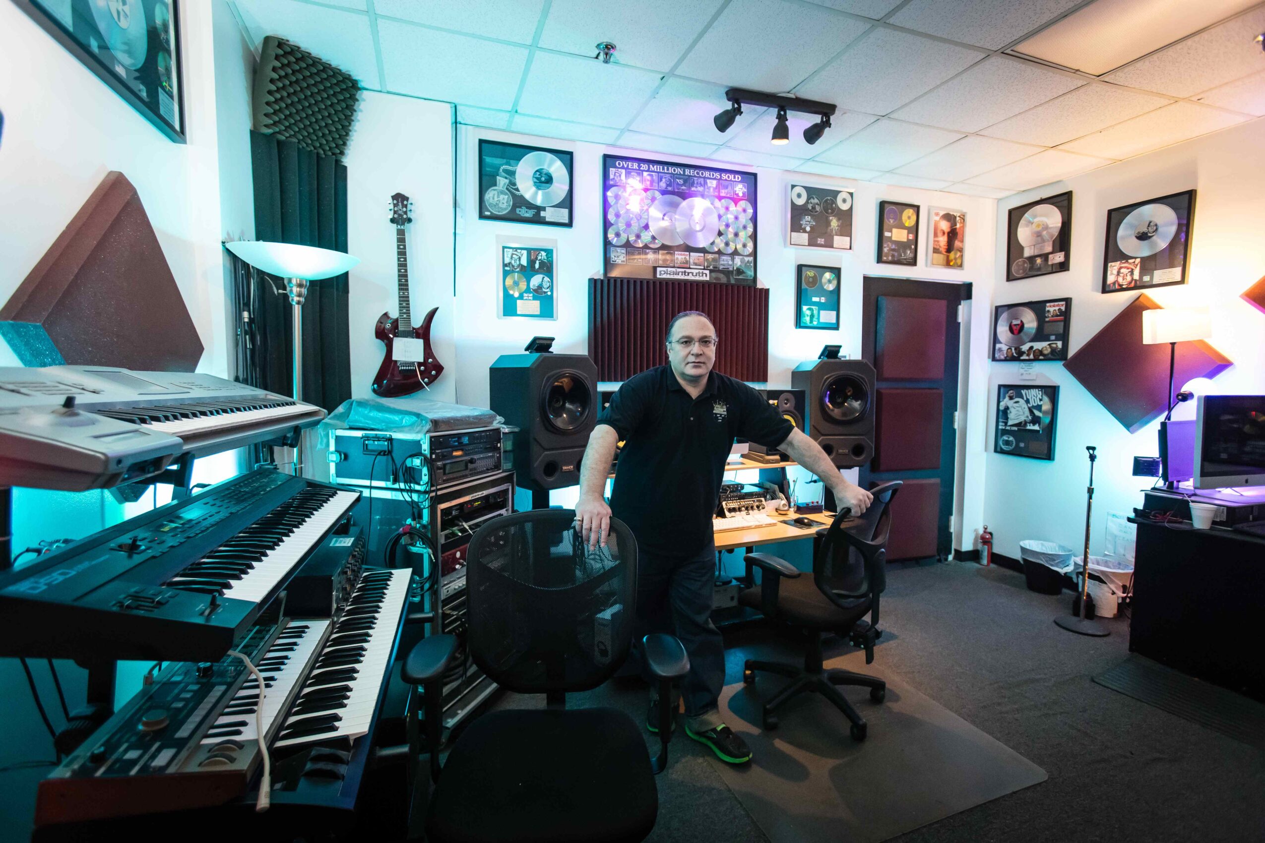 Person sitting in a music studio surrounded by instruments and framed records on the wall.