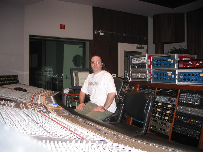 Person sitting in a recording studio with audio equipment and mixing console, facing camera, wearing a white shirt.