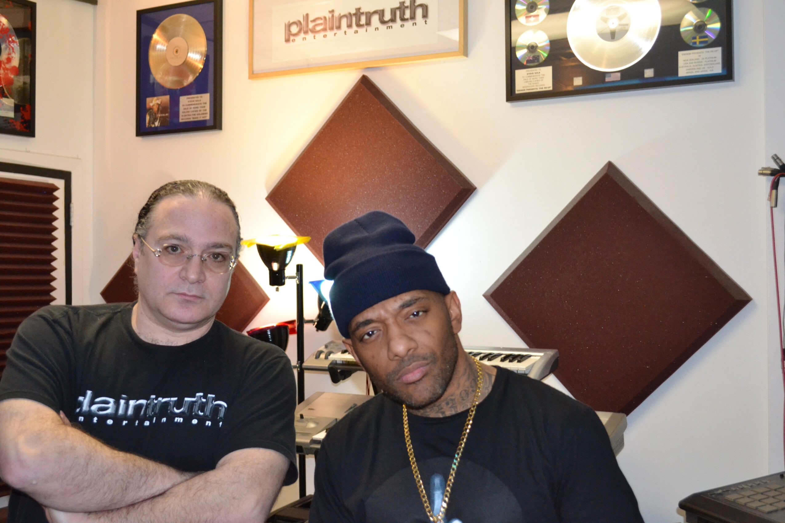 Two men sit in a room with framed gold records and soundproofing panels on the wall behind them.