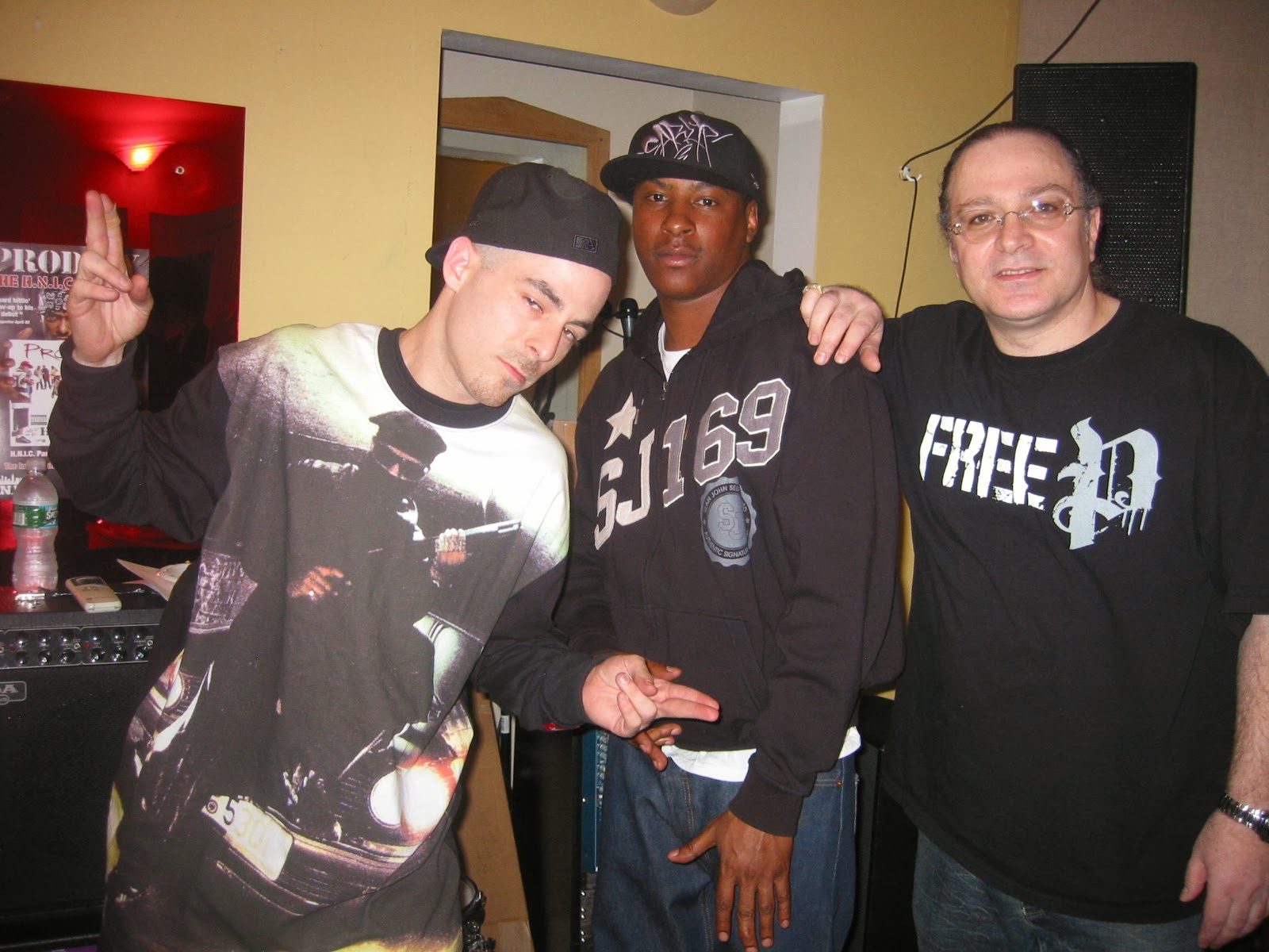 Three men standing together indoors, one with a black cap, another with a black hoodie, and the third with glasses, smiling.