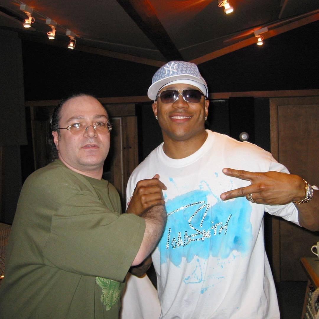 Two men pose together indoors, one wearing glasses and a green shirt, the other in a white shirt, sunglasses, and a cap, making a peace sign.