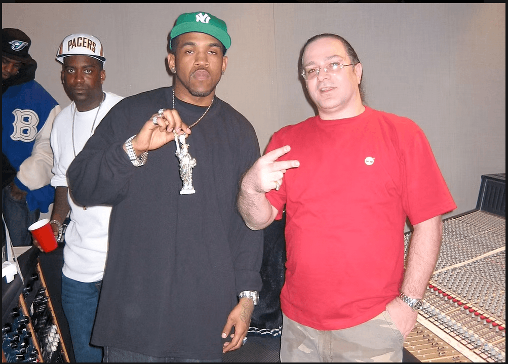 Three men standing indoors, one wearing a green cap and jewelry, another in a red shirt, and a third in a white cap and jacket.