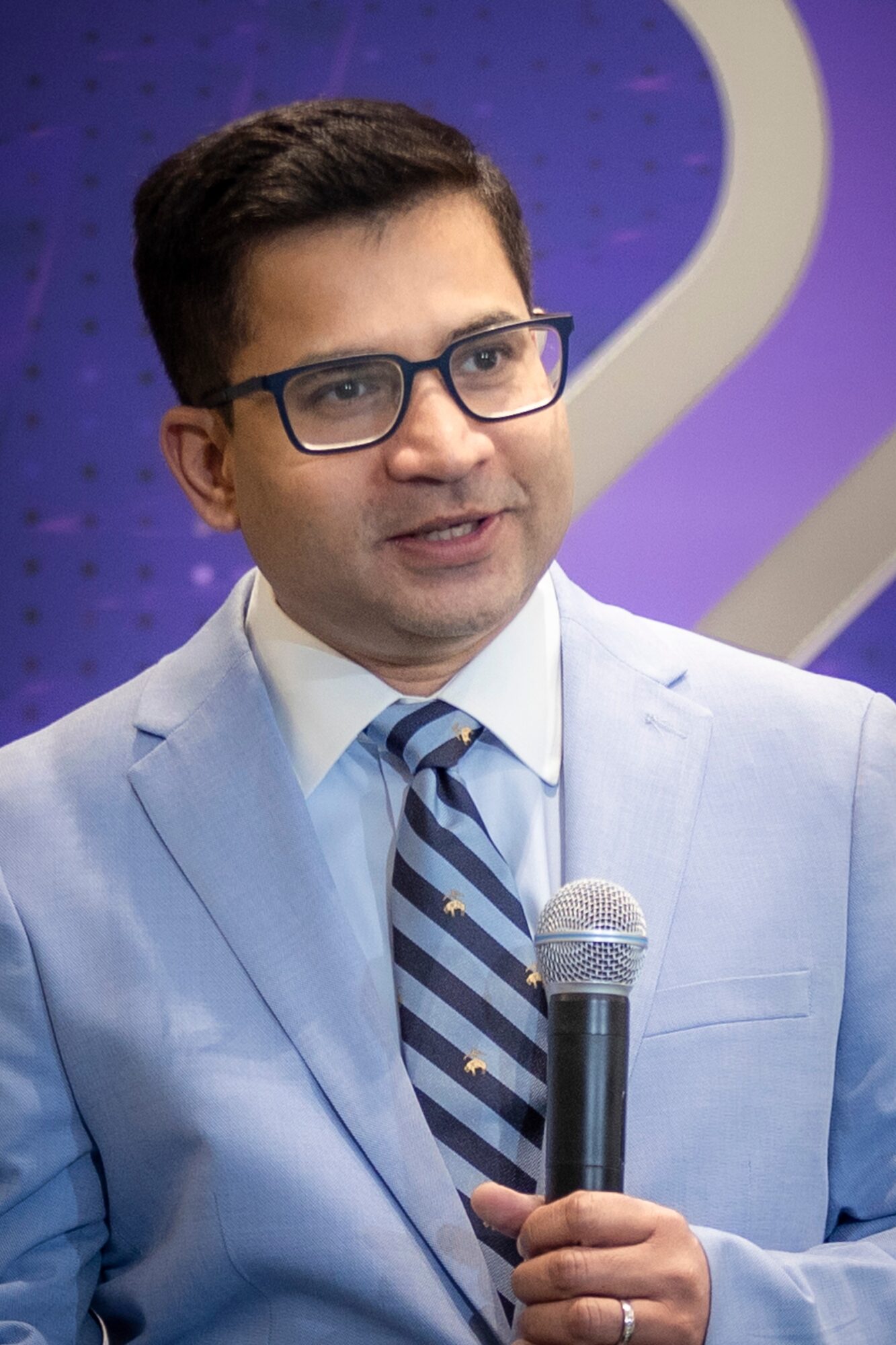 Man in glasses and light blue suit holding a microphone, speaking at an event, with a purple background.