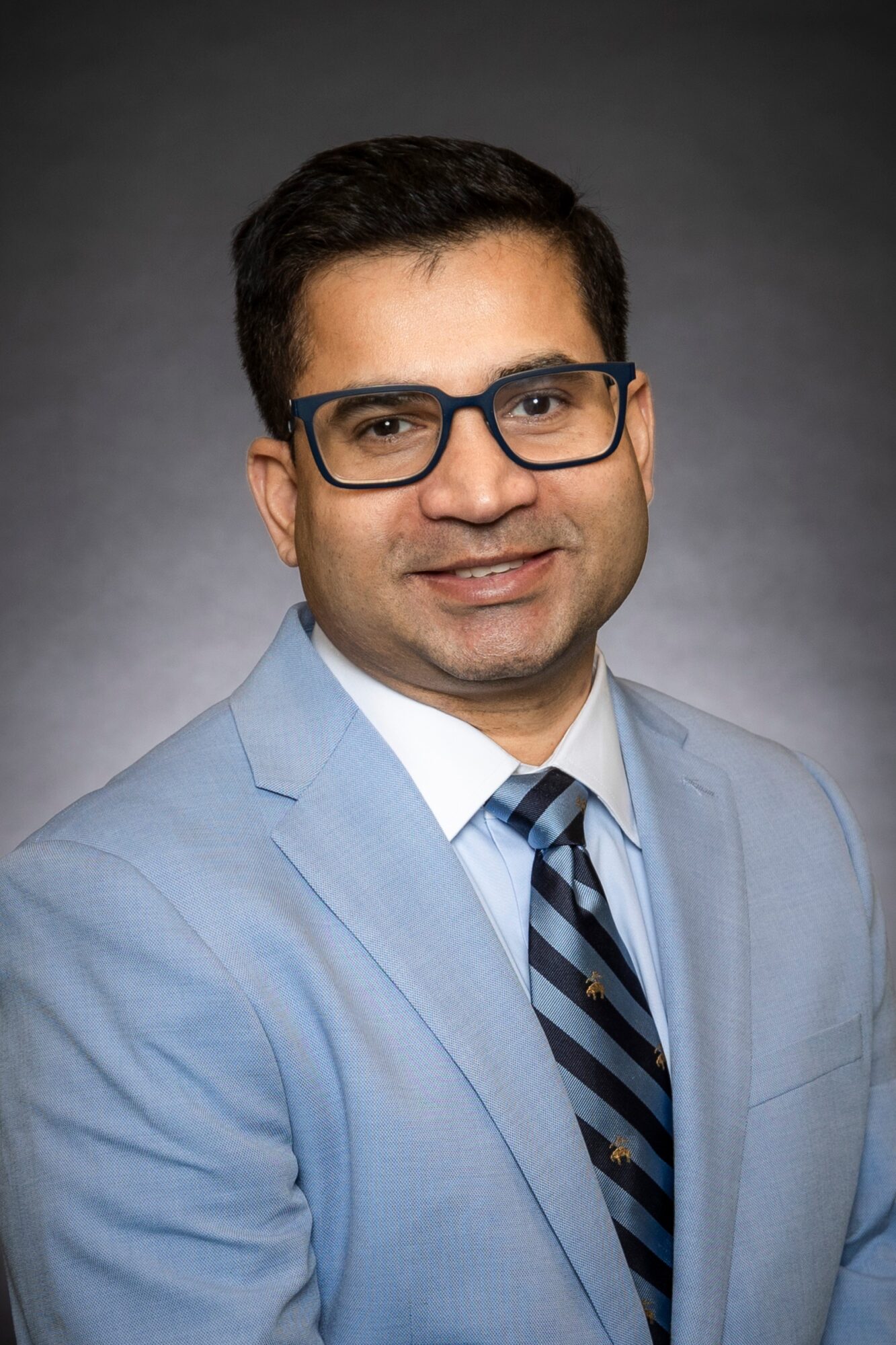 Portrait of a man wearing glasses, a light blue suit, white shirt, and striped tie, smiling against a dark background.