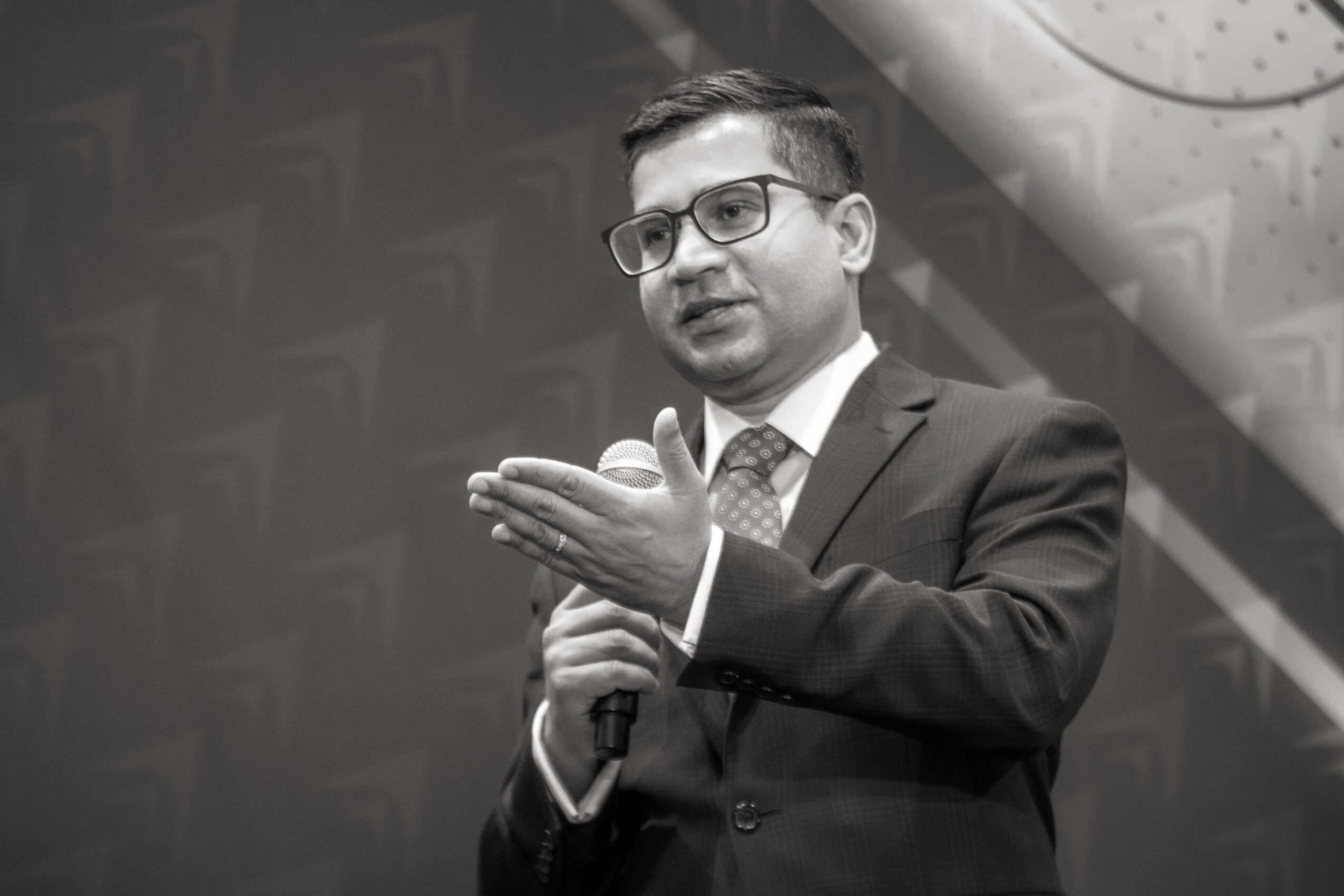 Man in suit and glasses speaking into a microphone, gesturing with his hand, in black and white.