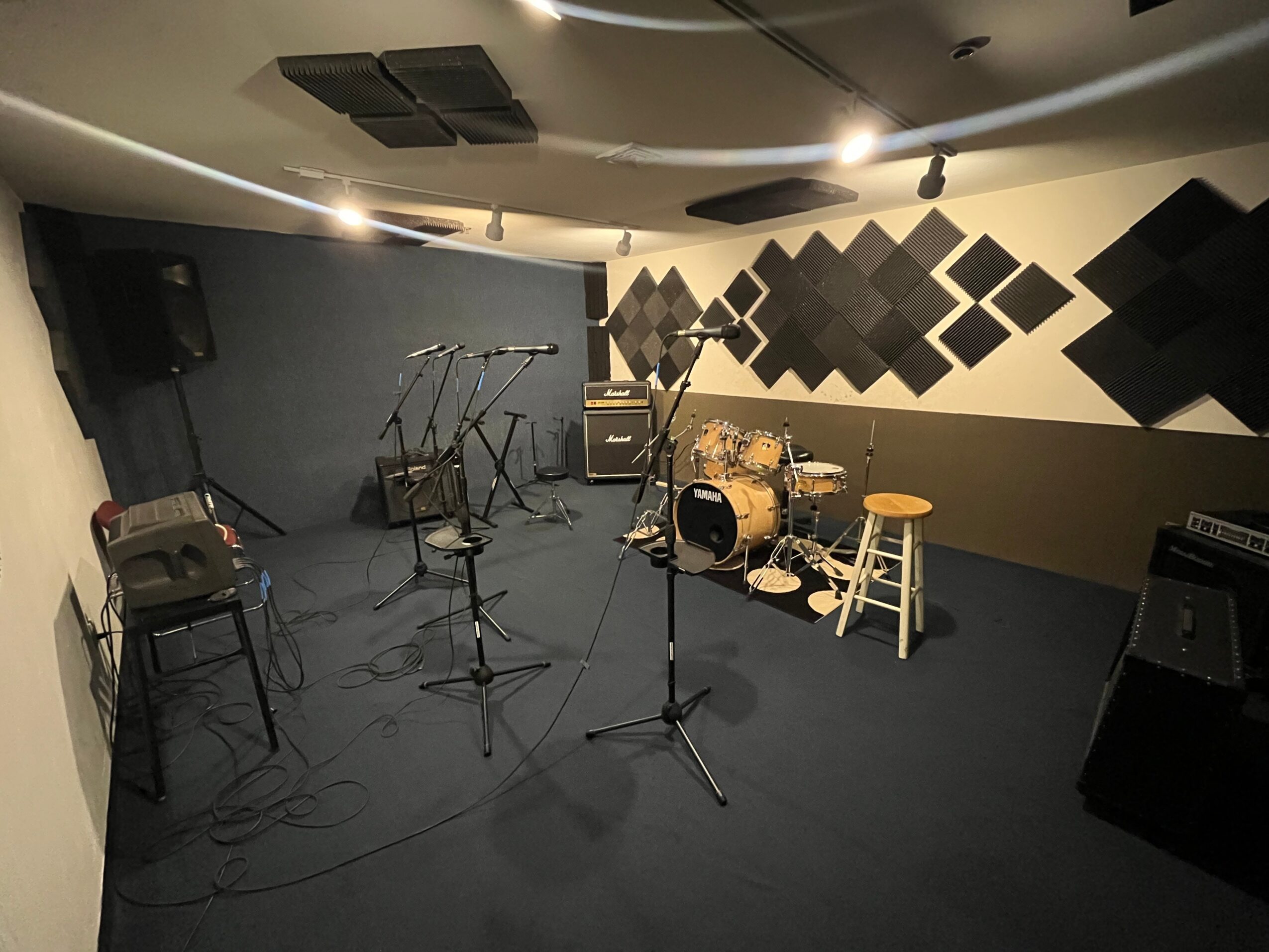Music rehearsal room with drums, microphones, chairs, and sound equipment, decorated with black acoustic panels on the wall.