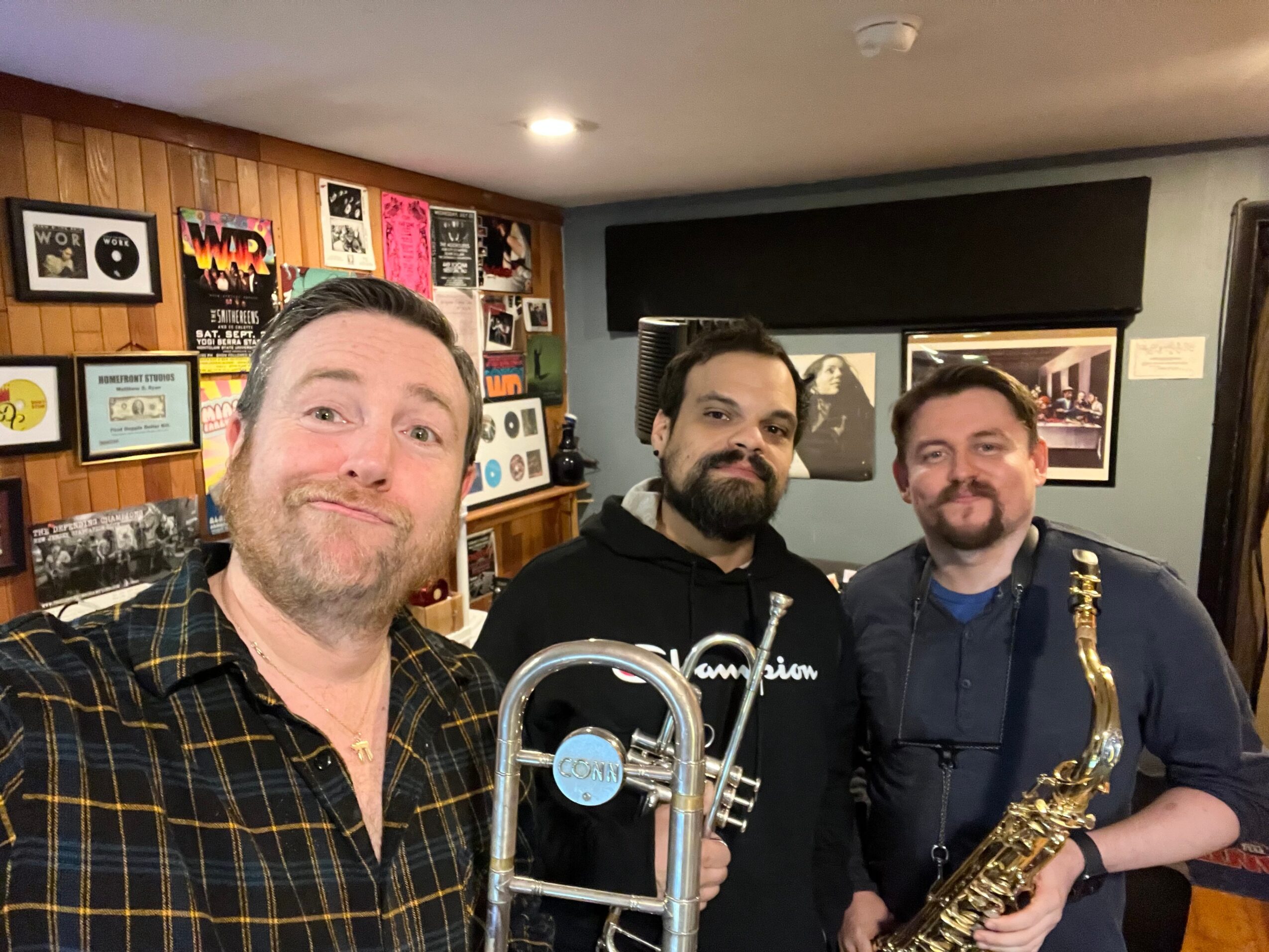Three men posing indoors, one holding a trombone and a saxophone, with a wall of framed pictures behind them.