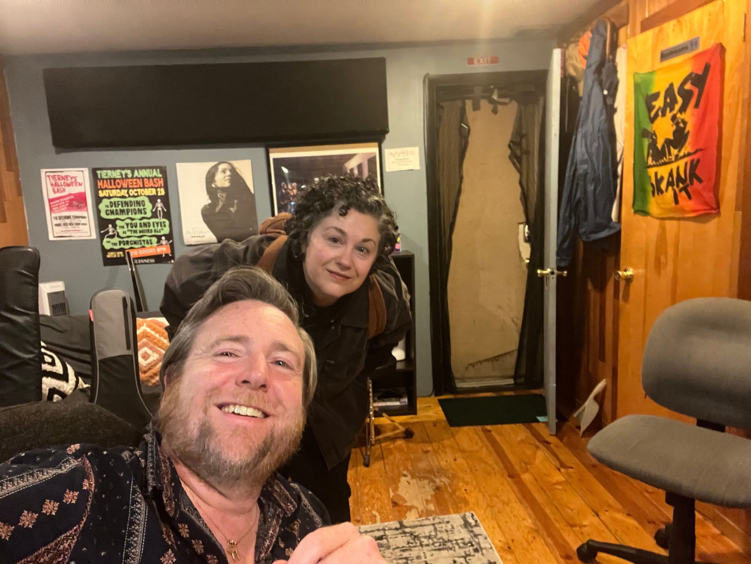Two people smiling and taking a selfie in a room with wooden walls and various objects, including a computer monitor and framed pictures.