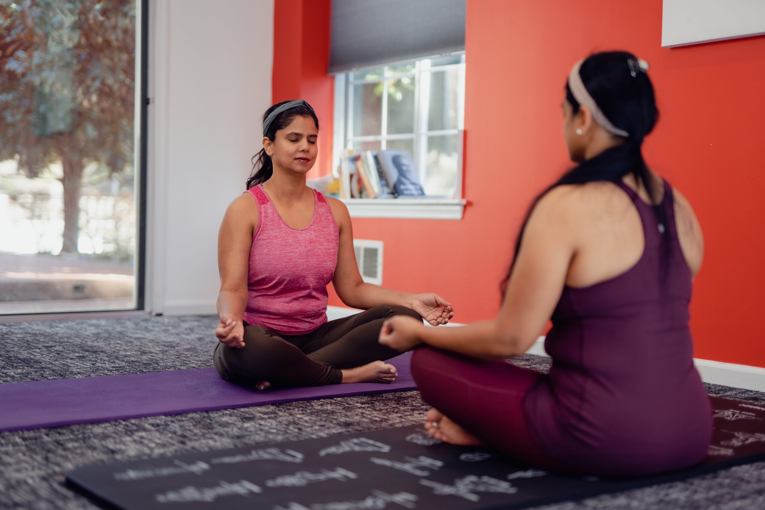 Two women practicing yoga indoors, sitting cross-legged on mats, facing each other with eyes closed, in a room with red walls and a window.