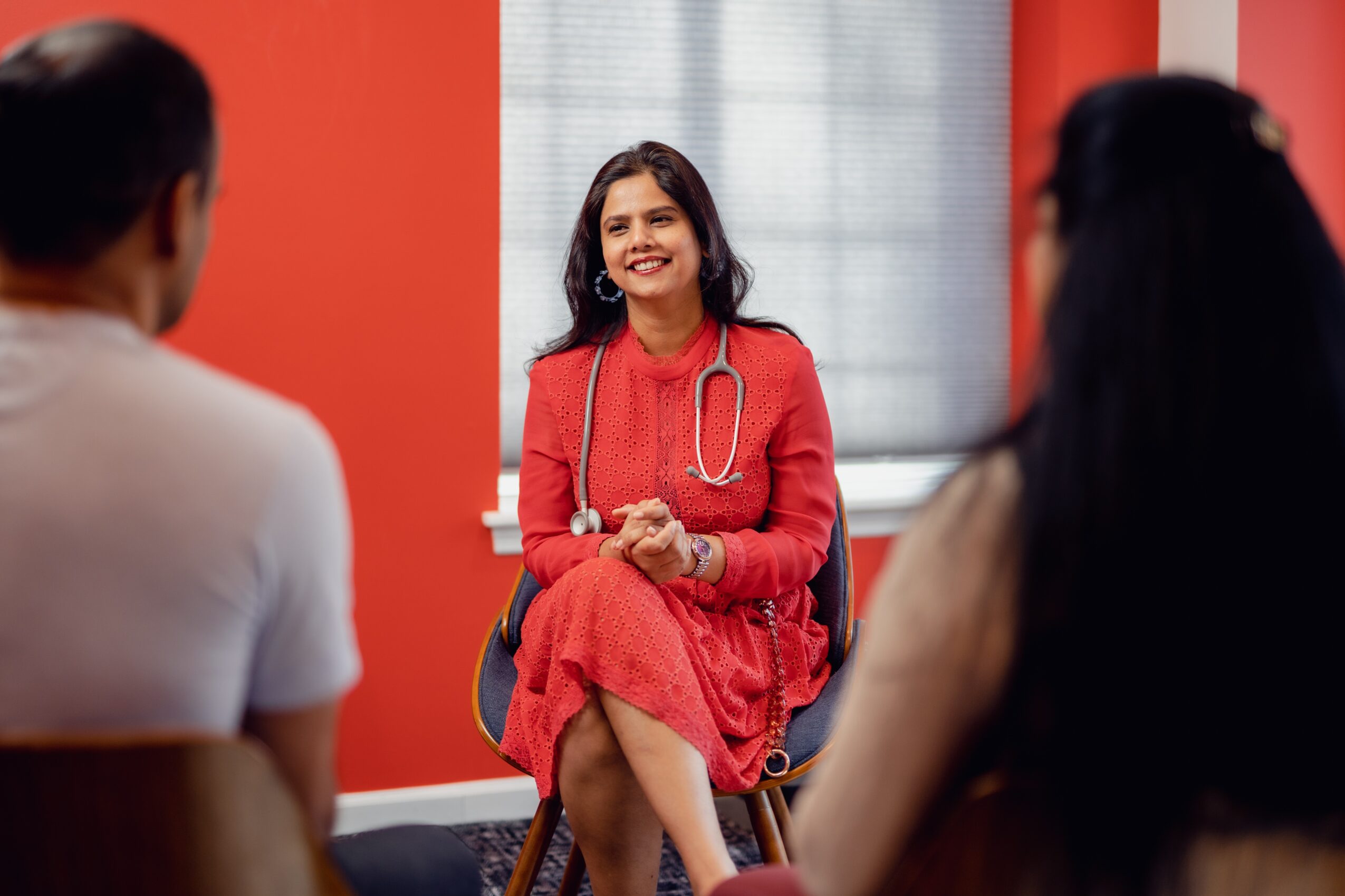 Young woman in red dress with stethoscope smiling, sitting in a room with two people facing her, red walls, window behind.