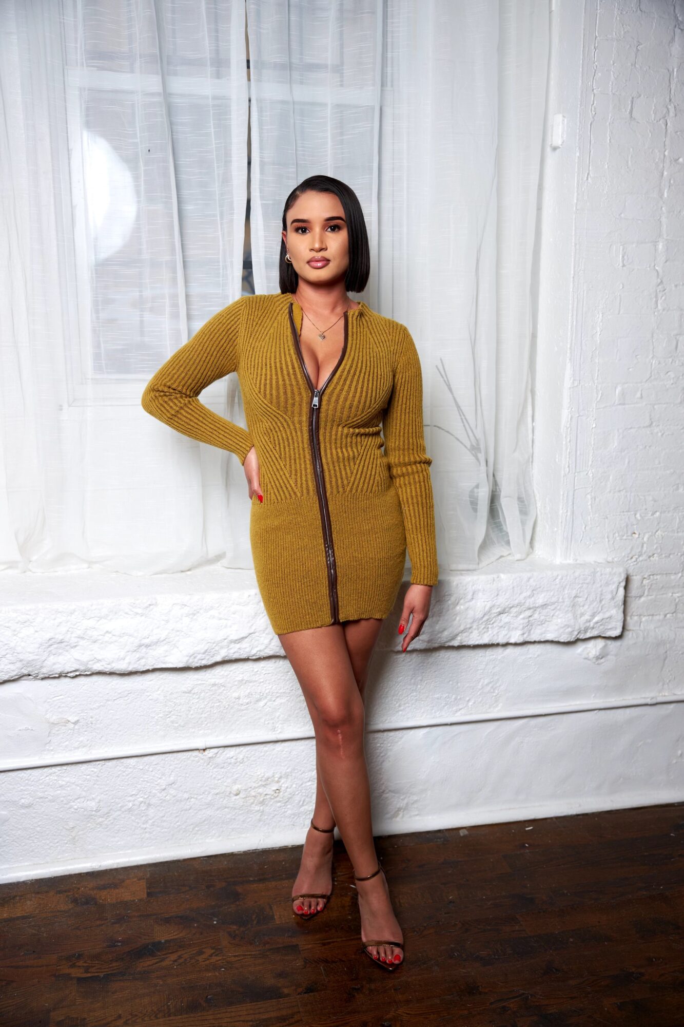 Woman in mustard yellow dress with zipper, standing indoors against white wall and window, with one hand on hip, looking at camera.
