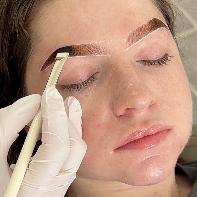 Person with closed eyes has eyebrows being shaped with a tool, wearing gloves, close-up of face, natural skin, makeup, and eyebrows.