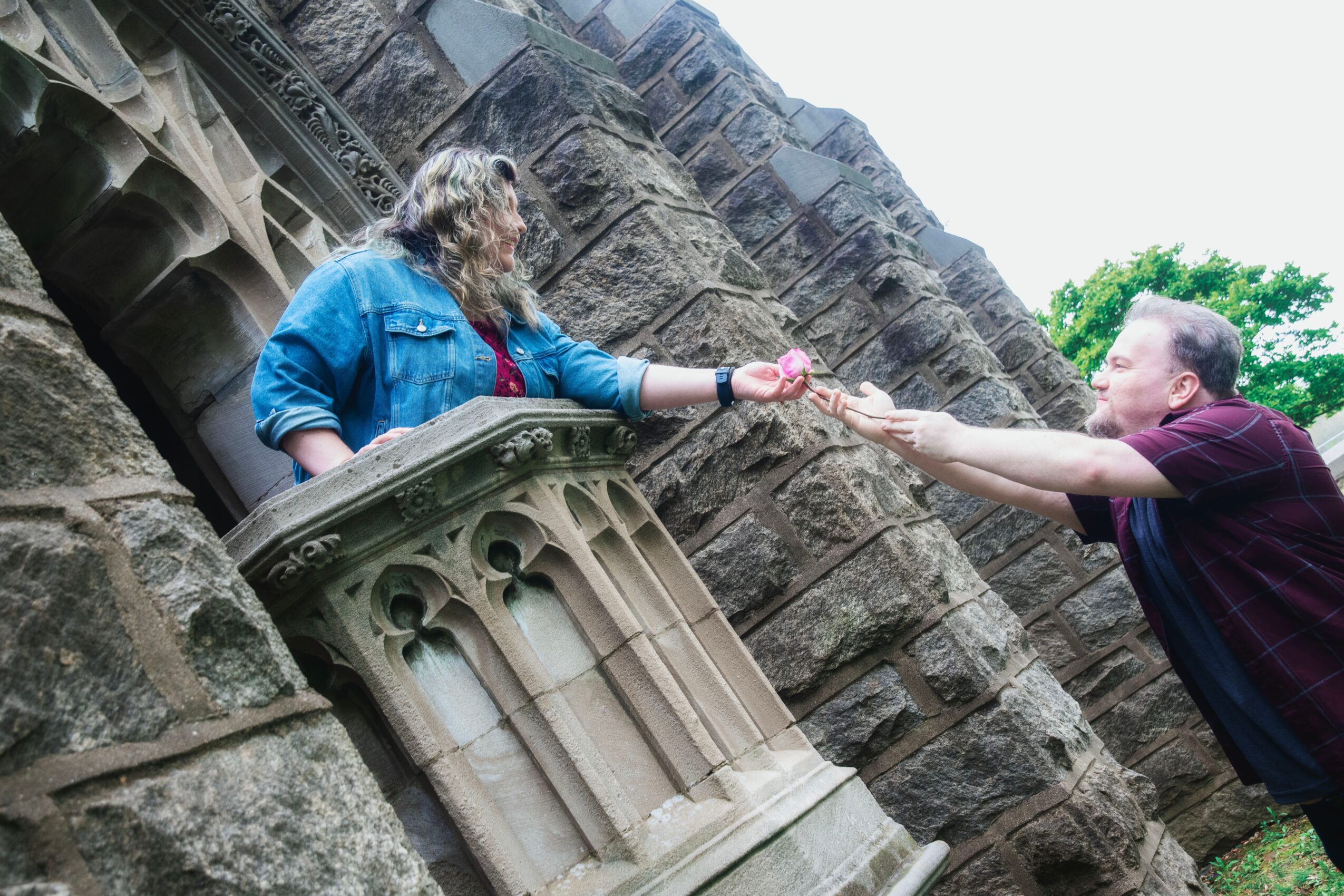 Two people exchange an object in front of a stone structure with arches, outdoors with trees in the background.