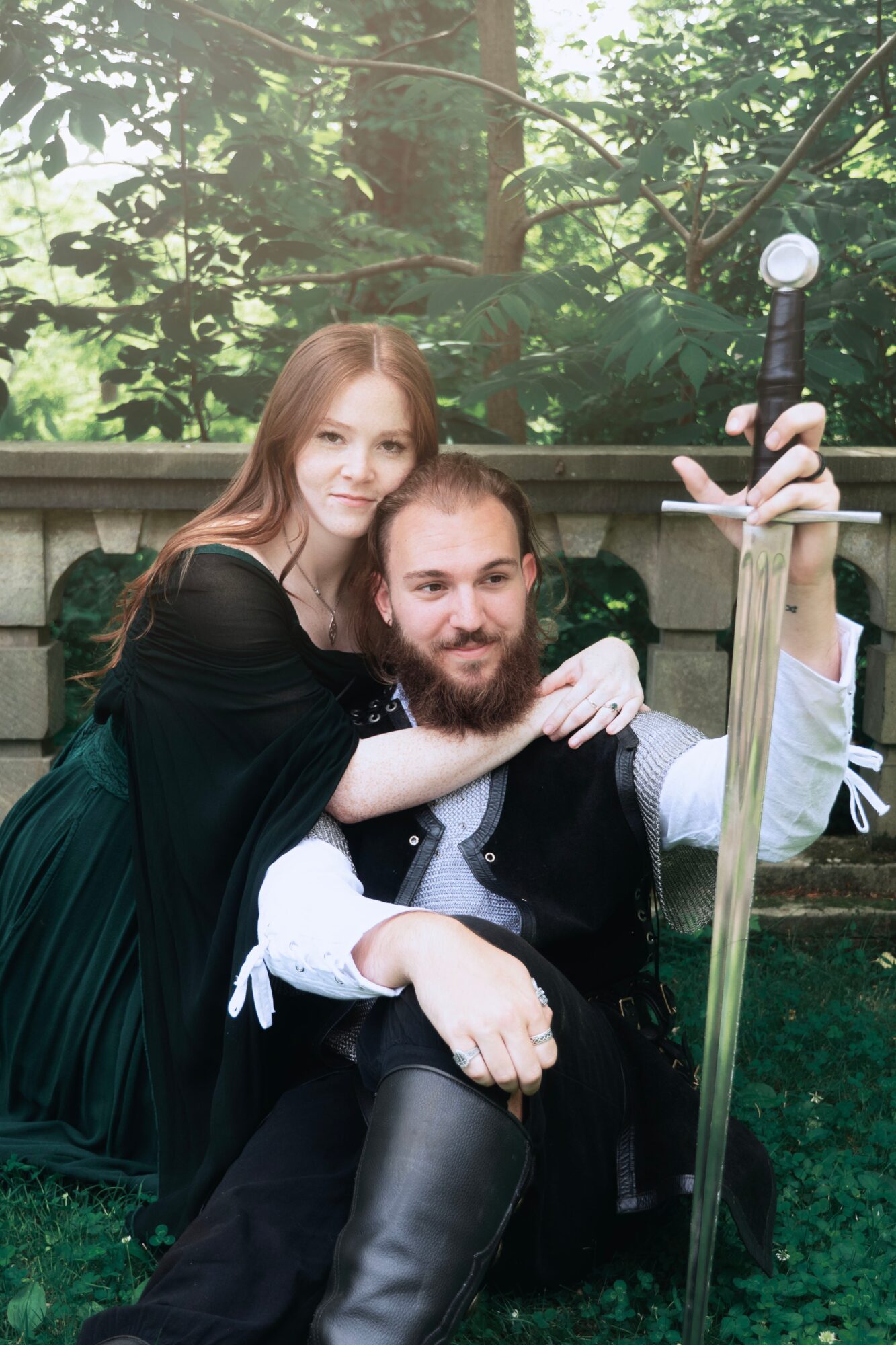 Two people dressed in medieval costumes pose outdoors, with a wooden fence and greenery in the background.