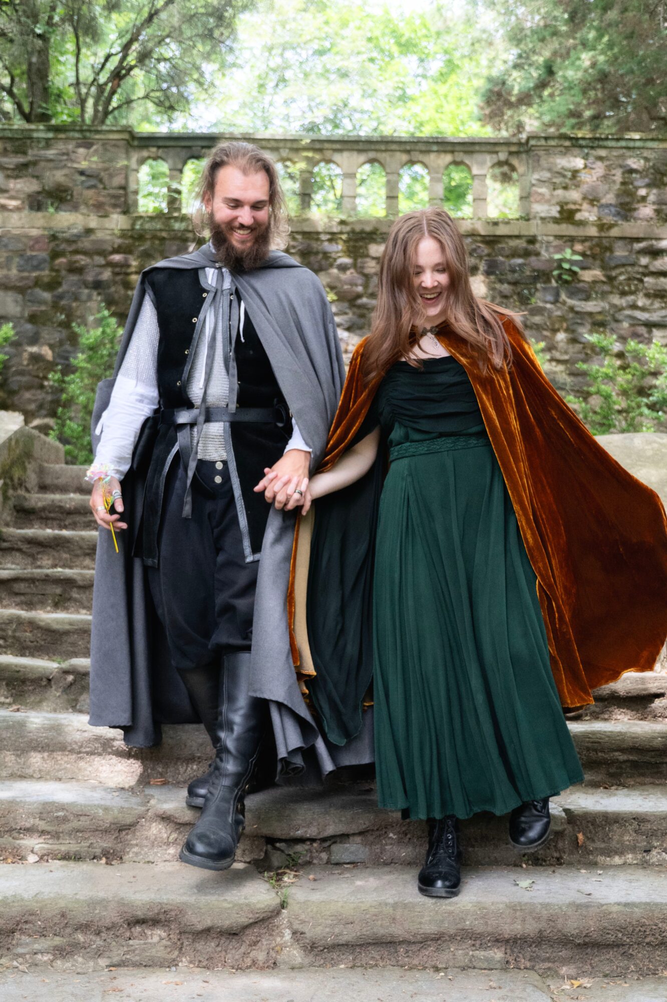 Two people in medieval costumes standing on stone steps outdoors, with a stone wall and trees in the background.