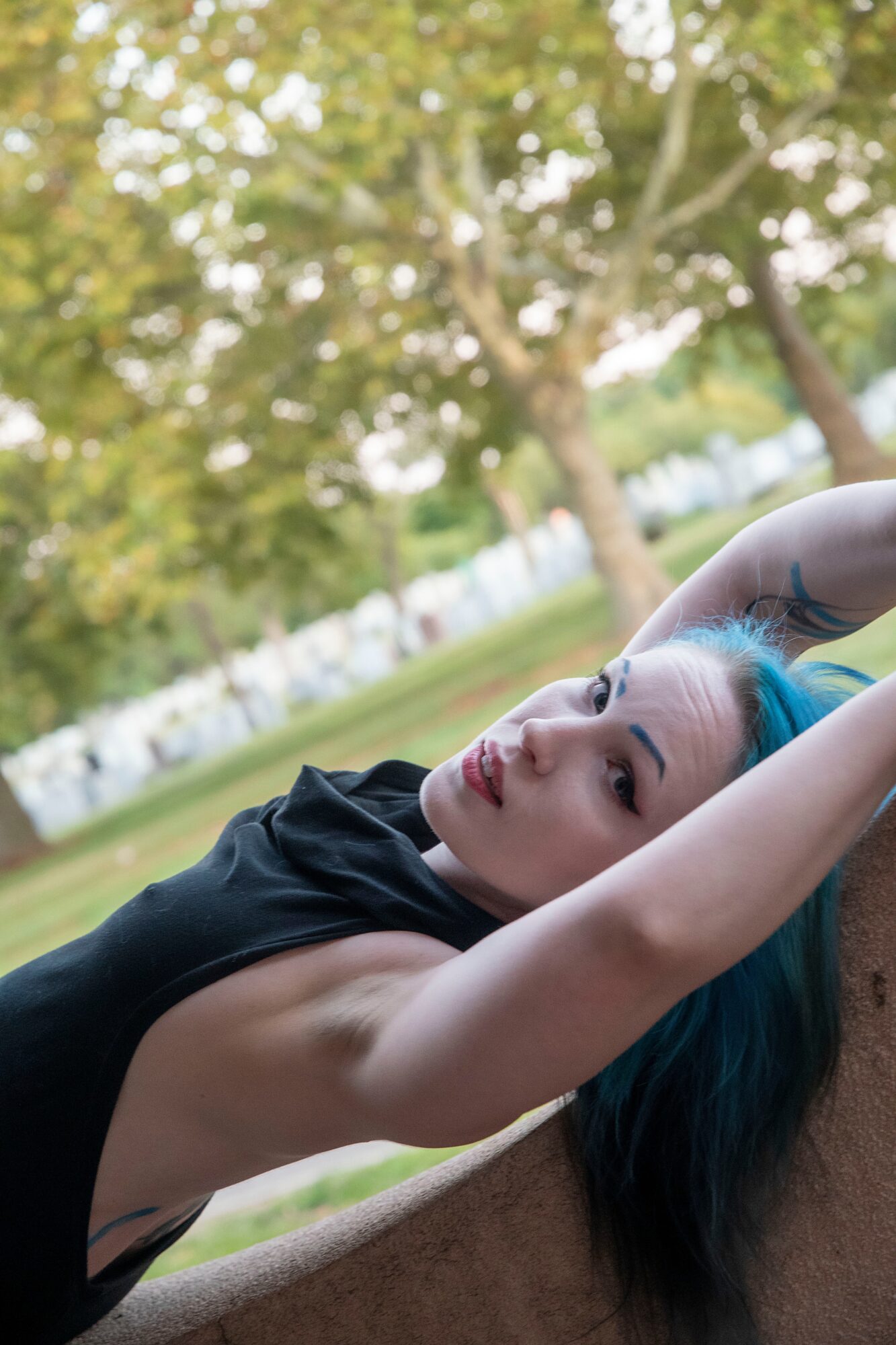 Young woman with blue hair lying outdoors on grass, arms behind head, trees in background, relaxed expression.